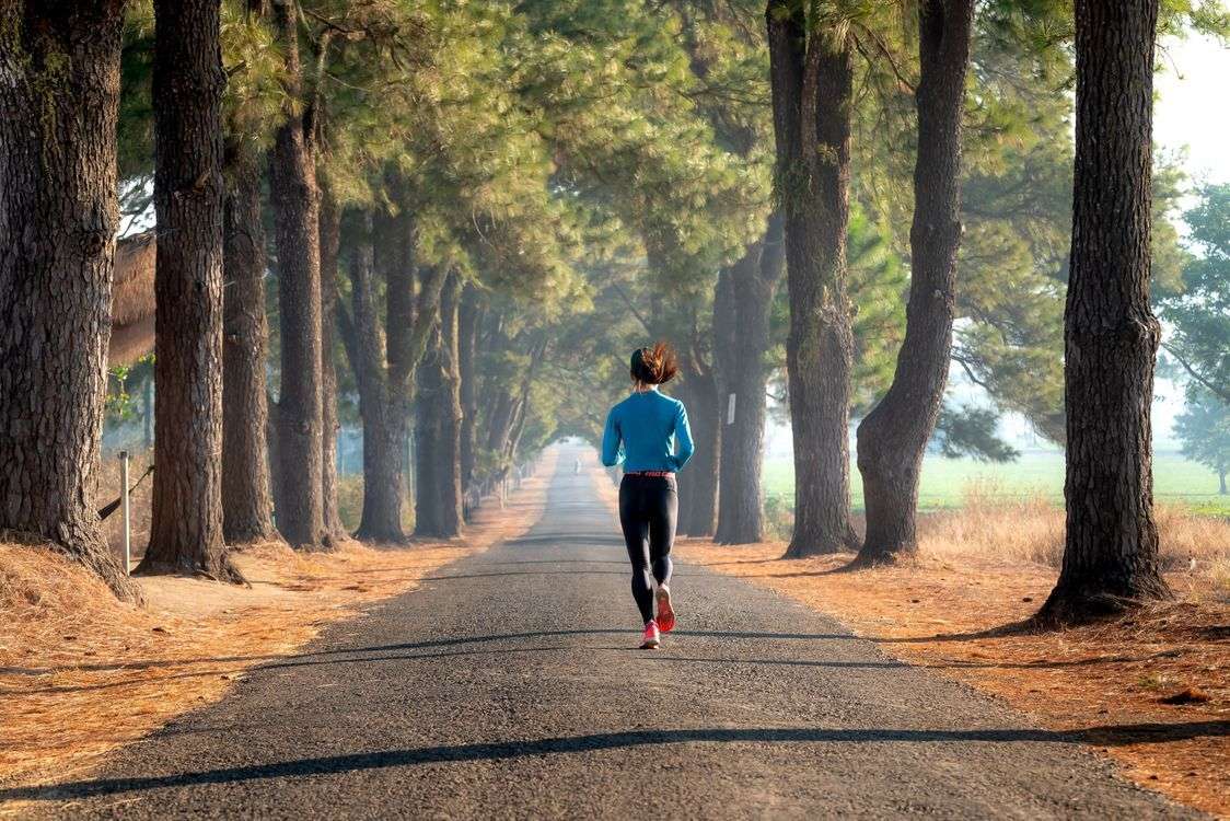 Woman running on a tree-lined road at sunrise, an example of the aerobic exercise that helps lower resting heart rate naturally.