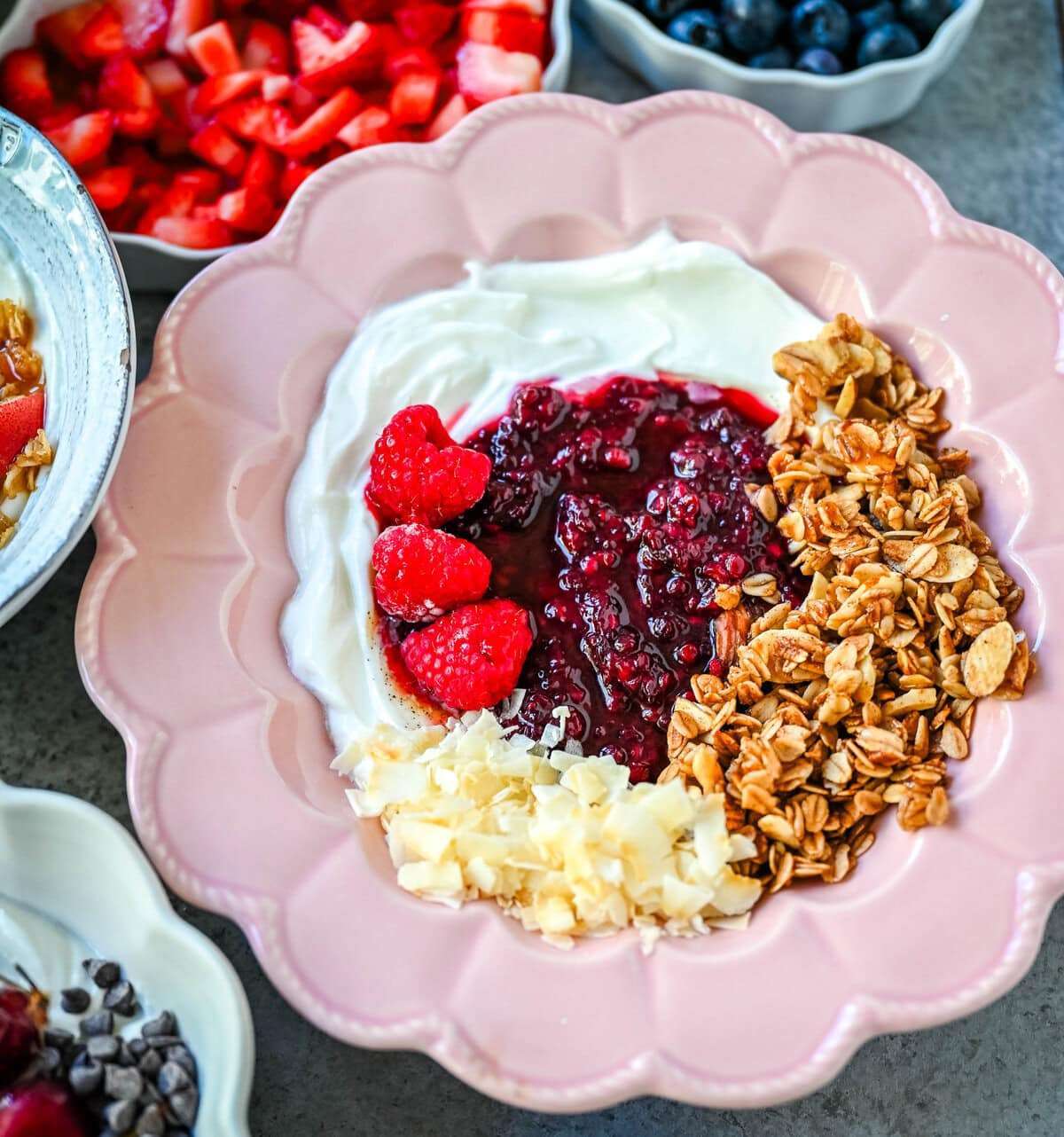 A pink bowl of Greek yogurt topped with raspberry compote, fresh raspberries, granola, and shredded coconut, with small bowls of diced strawberries, blueberries, and chocolate chips nearby.