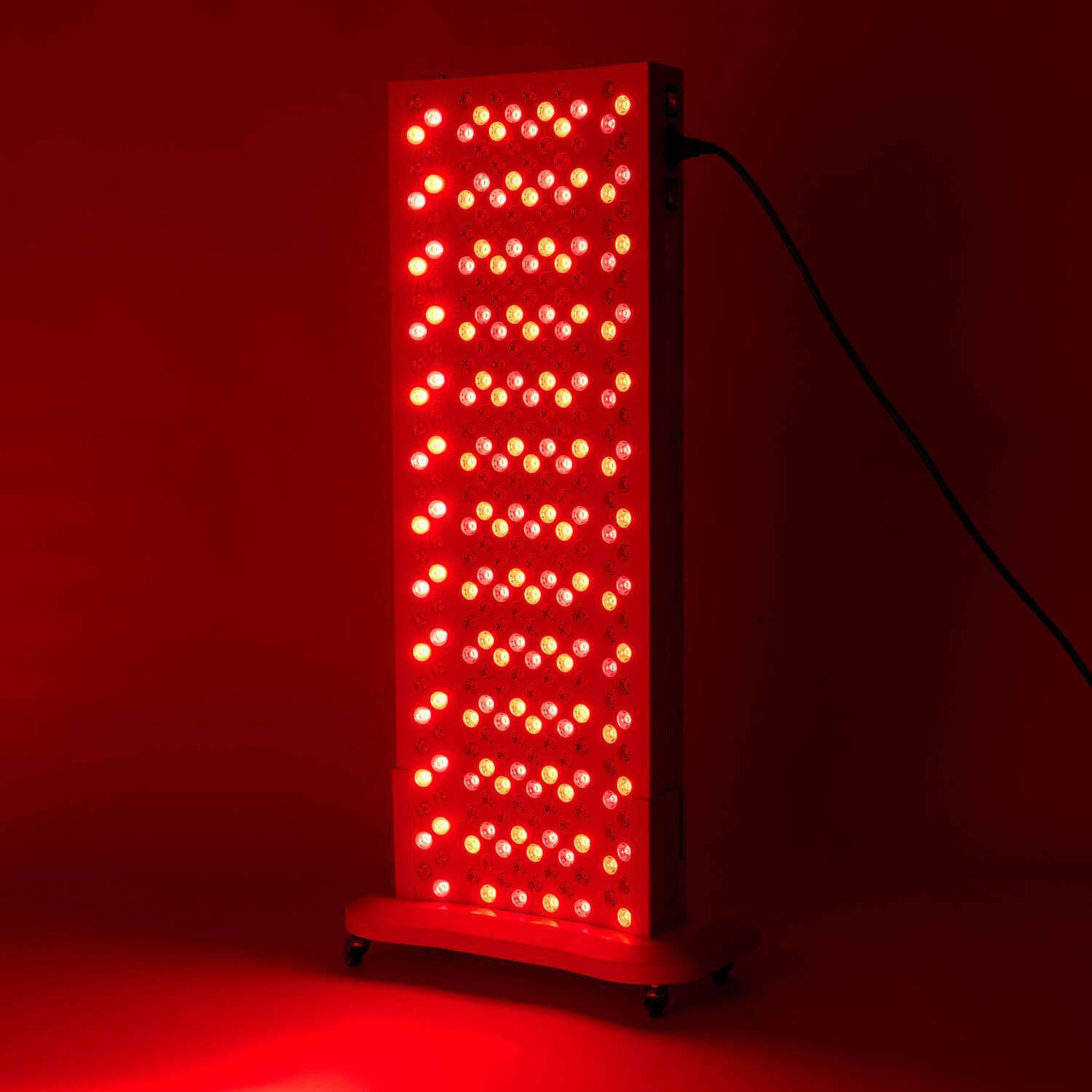 A full-body red light therapy panel device with rows of glowing red and near-infrared LED bulbs mounted on a rolling stand against a dark background.