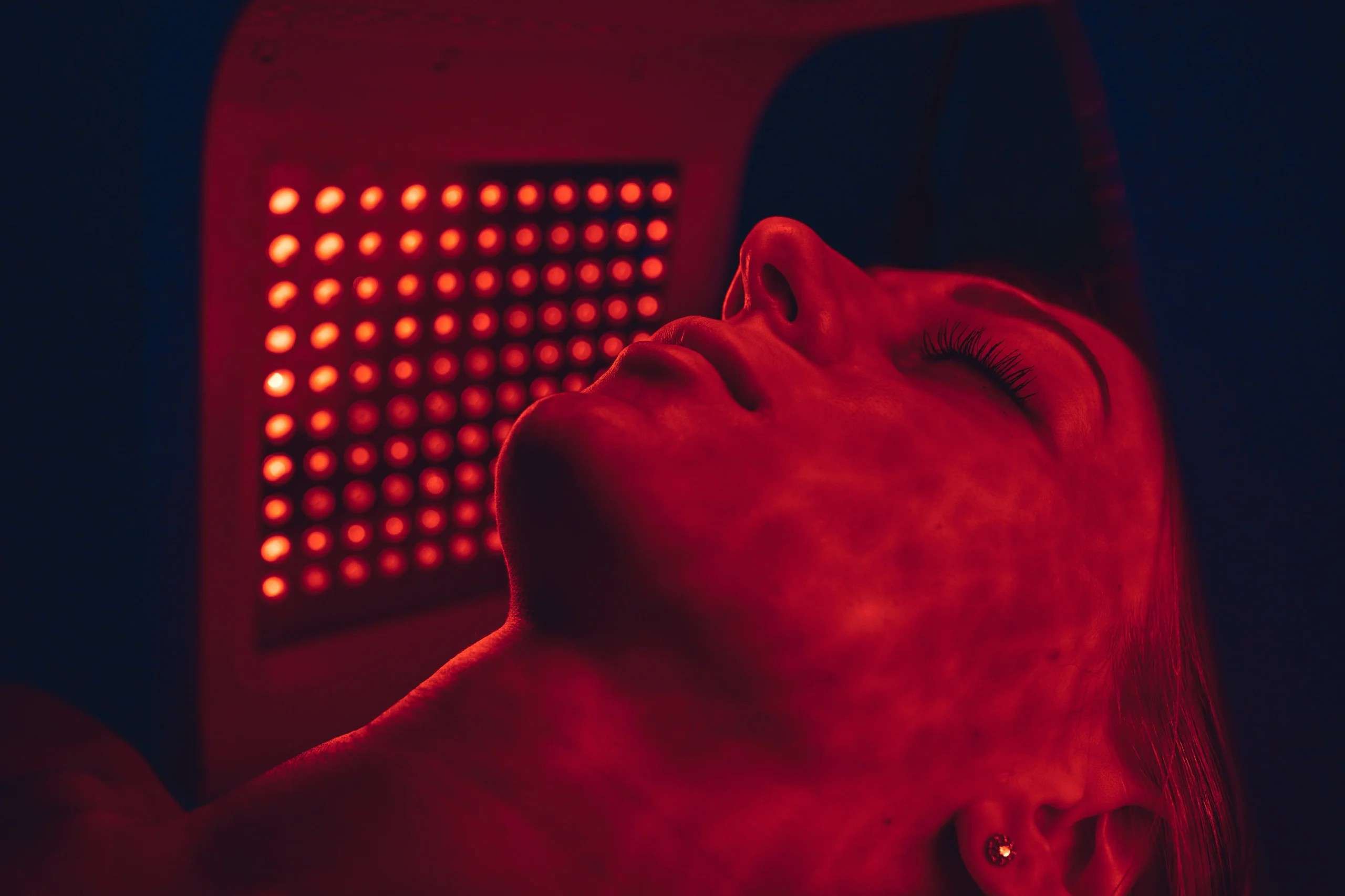 Close-up of a woman's face bathed in warm red light from a red light therapy panel during a treatment session.