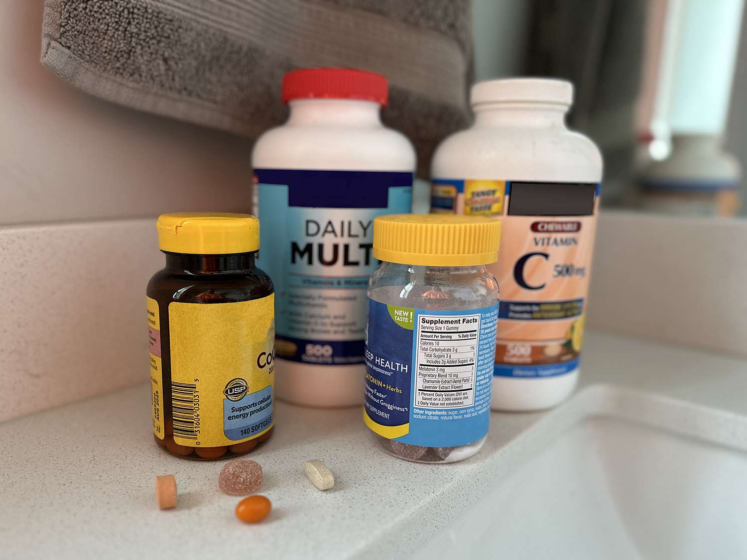 Several supplement bottles including a daily multivitamin, vitamin C, and sleep gummies arranged on a bathroom counter with loose pills in front.