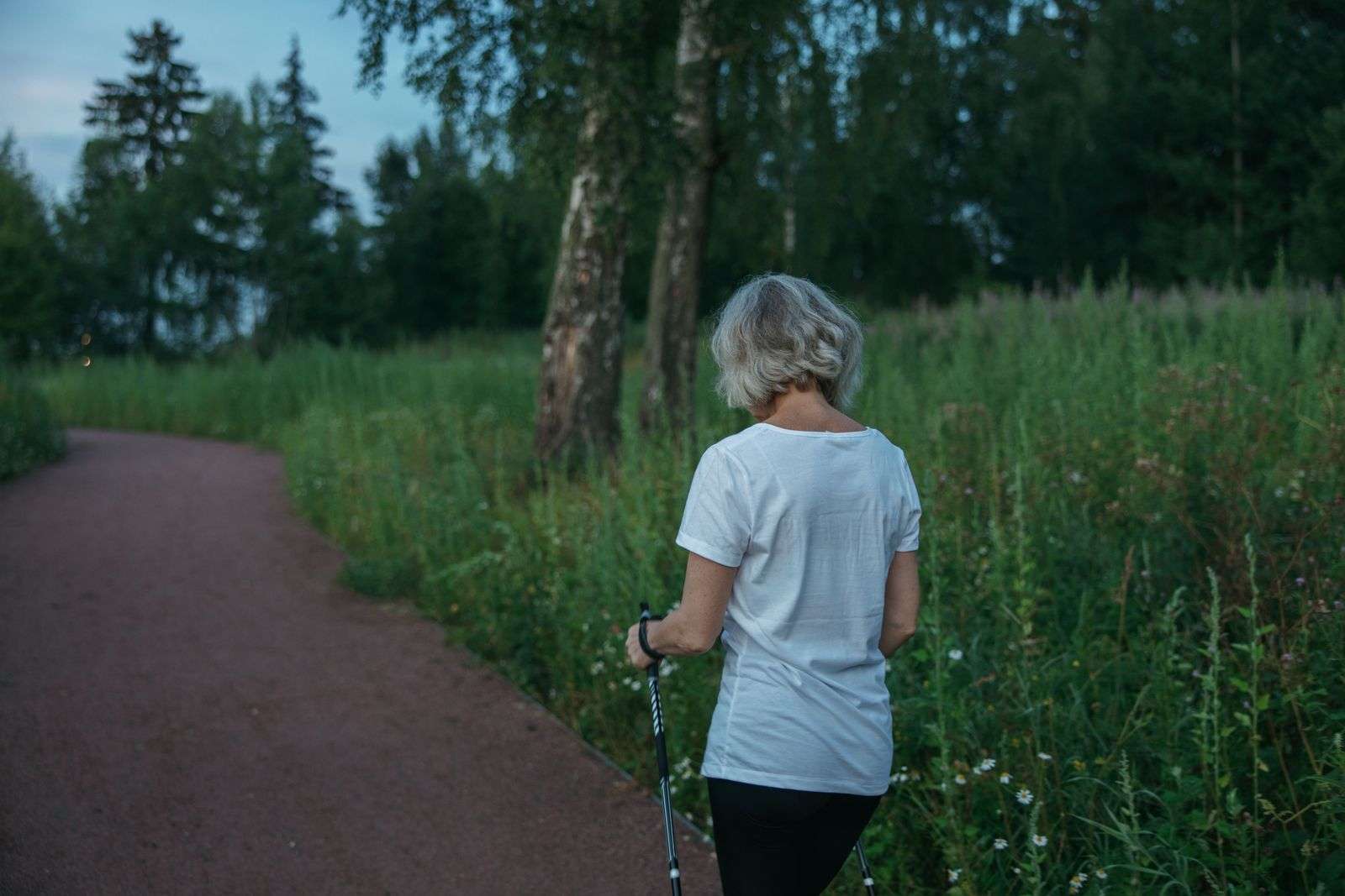 Elderly woman practicing Nordic walking with poles on a park trail at dusk, an active-aging example of walking for longevity